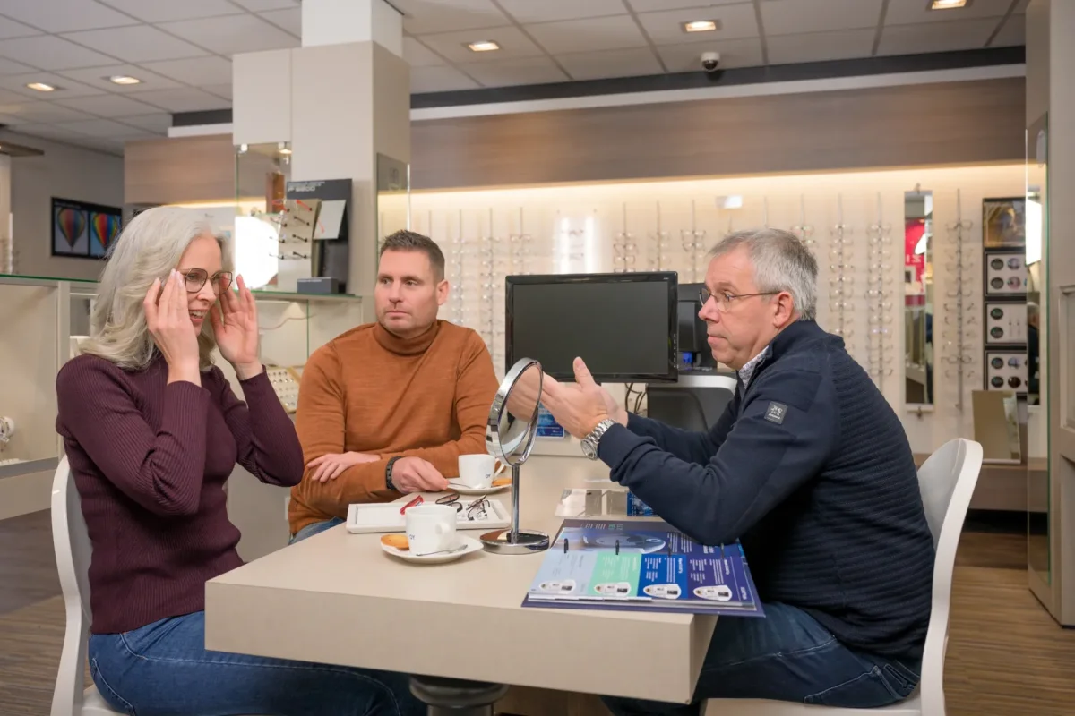 Opticien Cris in gesprek aan tafel met een klant die haar hand tegen haar hoofd houdt