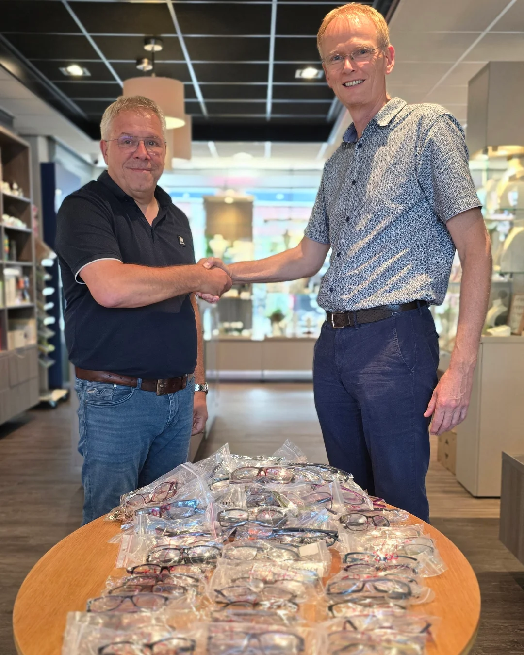 Opticien Cris geeft huisarts Arntzen een hand, 300 brillen voor Zambia Opticien Cris van Von Oy (links) geeft huisarts Arntzen een hand, met de brillen voor Zambia op tafel in de winkel
